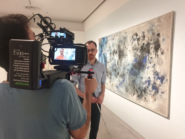A cameraman films an interview with an expert in a museum with a painting in the background